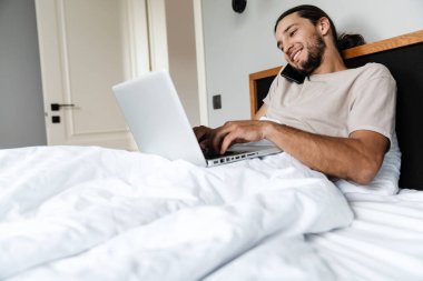 Gülümseyen genç adam sabah yatağında yatarken cep telefonu ve dizüstü bilgisayar kullanıyor.