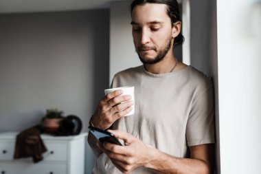 Evde elinde fincanla cep telefonu kullanan çekici bir genç.