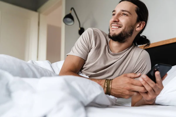 Smiling young man using mobile phone while laying in bed in the morning