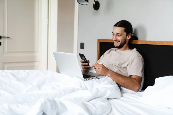 Joven Sonriente Usando Teléfono Móvil Computadora Portátil Mientras Está Acostado —  Fotos de Stock