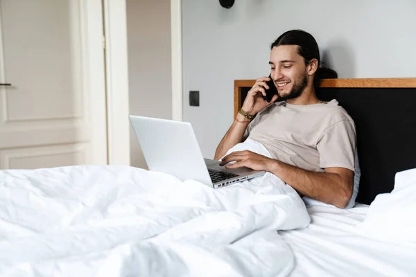 Uśmiechnięty Młodzieniec Korzystający Telefonu Komórkowego Laptopa Podczas Leżenia Rano Łóżku — Zdjęcie stockowe