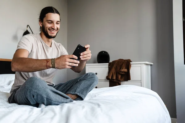 Joven Atractivo Sonriente Hombre Caucásico Sentado Cama Dormitorio Uso Teléfono —  Fotos de Stock