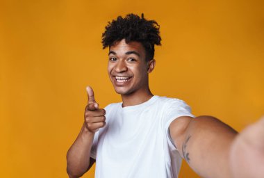 Sarı duvardan izole edilmiş selfie çekerken kamerayı işaret eden mutlu Afrikalı Amerikalı bir adam.