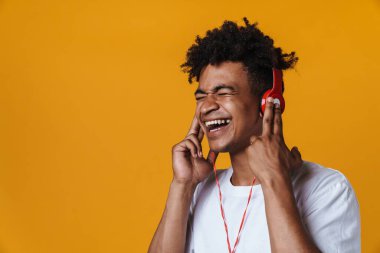 Heyecanlı Afrikalı Amerikalı bir adam kulaklıklı müzik dinliyor ve sarı duvarda izole edilmiş şarkı söylüyor.