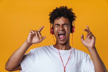 Heyecanlı Afrikalı Amerikalı bir adam kulaklıklı müzik dinliyor ve sarı duvarda izole edilmiş şarkı söylüyor.