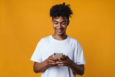 Sarı duvardan izole edilmiş gülümseyen ve cep telefonu kullanan mutlu Afro-Amerikan bir adam.