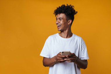 Sarı duvardan izole edilmiş gülümseyen ve cep telefonu kullanan mutlu Afro-Amerikan bir adam.