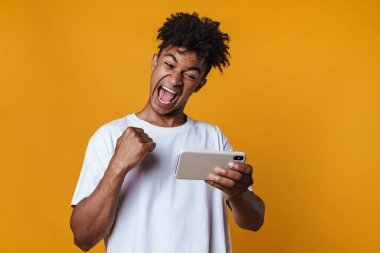Afro-Amerikan bir adamın sarı duvarda izole edilmiş bir şekilde kazanan bir jest yapması ve cep telefonuyla oyun oynaması.