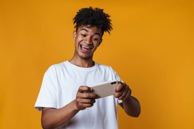 Sarı duvarda izole edilmiş cep telefonuyla online oyun oynayan heyecanlı Afrikalı Amerikalı adamın görüntüsü