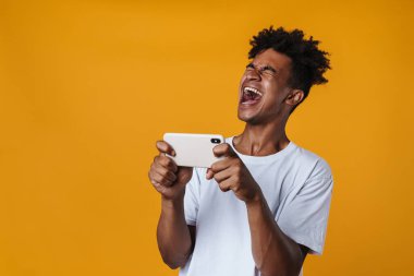 Sarı duvarda izole edilmiş cep telefonuyla online oyun oynayan heyecanlı Afrikalı Amerikalı adamın görüntüsü