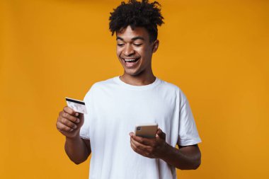 Sarı duvardan izole edilmiş kredi kartı ve cep telefonu tutan mutlu Afrikalı Amerikalı bir adam.
