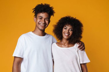 Sarı duvarda izole edilmiş kameraya sarılıp gülümseyen mutlu Afro-Amerikan çifti.