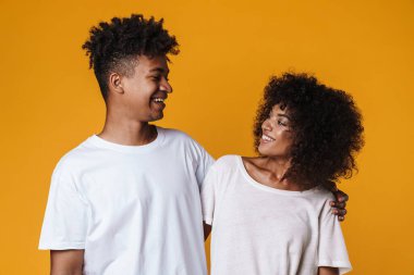 Sarı duvarda izole edilmiş kameraya sarılıp gülümseyen mutlu Afro-Amerikan çifti.
