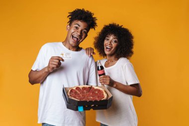 Kredi kartıyla pizza yiyen ve sarı duvardan izole edilmiş gazoz içen Afro-Amerikan çifti.