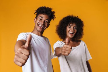 Heyecanlı Afrikalı Amerikalı bir çiftin sarı duvarın üzerinde gülümsemesi ve başparmaklarını göstermesi.