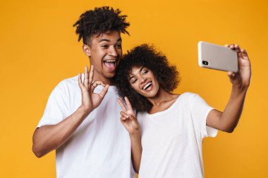 Sarı duvarda izole edilmiş akıllı telefonlardan selfie çekerken barış işareti yapan Afro-Amerikan çifti.
