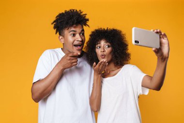 Afro-Amerikan çiftin, sarı duvarda izole edilmiş akıllı telefondan selfie çekerken kamerayı işaret etmesi.