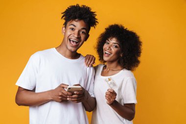 Gülümseyen Afrikalı Amerikalı bir çiftin görüntüsü. Kredi kartı ve akıllı telefon sarı duvarda izole edilmiş.