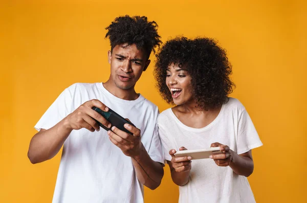 Photo of excited african american people playing online game on cellphones isolated over yellow background