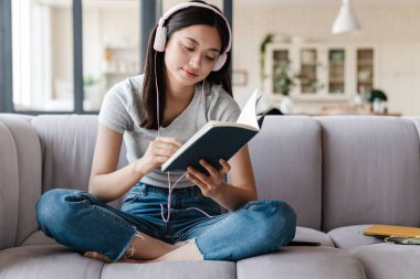 Evde kanepede otururken kulaklık takan ve notlar yazan odaklanmış Asyalı kadın görüntüsü