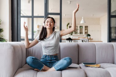 Evdeki kanepede otururken kulaklıkları açık müzik dinleyen mutlu Asyalı kadın resmi.