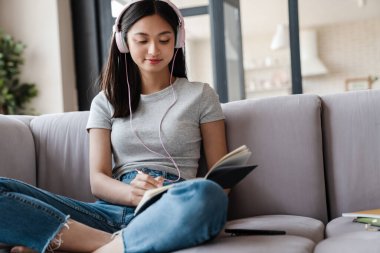 Evdeki kanepede otururken kulaklıklı Asyalı bir kızın notlar yazması.