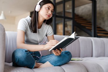 Evde kanepede otururken kulaklık takan ve notlar yazan odaklanmış Asyalı kadın görüntüsü