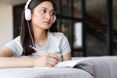 Evde kanepede otururken kulaklıklı Asyalı bir kız öğrencinin notlar yazması.