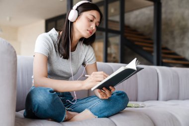 Evde kanepede otururken kulaklık takan ve notlar yazan odaklanmış Asyalı kadın görüntüsü