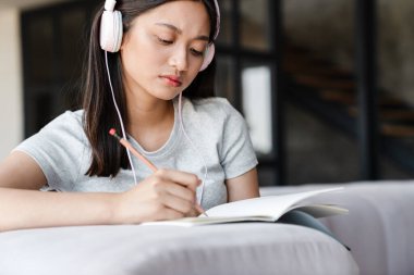 Evde kanepede otururken kulaklıklı Asyalı bir kız öğrencinin notlar yazması.