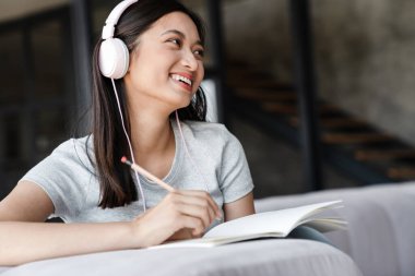 Evde kanepede otururken kulaklıkla notlar yazan Asyalı bir öğrenci resmi.