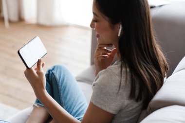 Evde koltukta otururken kulaklıkla cep telefonu kullanan Asyalı bir kadın resmi