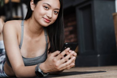 Spor kıyafetli neşeli Asyalı bir kızın evde yoga minderinde yatarken cep telefonu kullanması.