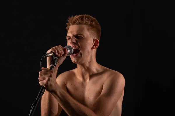 Topless guy sings punk rock in a microphone on a dark background