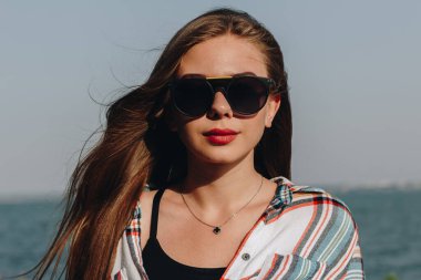 Sahilde güneş gözlüklü ve kırmızı dudaklı genç bir kızın portresi.