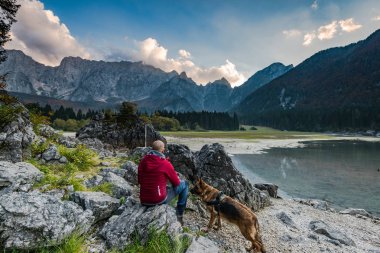 Adventure man with dog sitting at rock looking at alpine lake. clipart