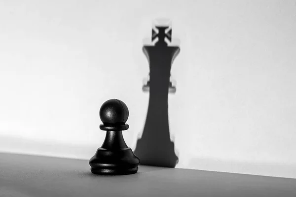 Chess pawn standing in a spotlight that make a shadow of king with darkness artistic conversion. Horizontal frame