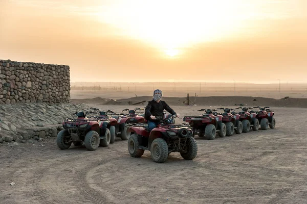 Un mec avec un mouchoir fermé sur le visage s'assoit sur un quad sur fond de soleil levant sur le désert — Photo