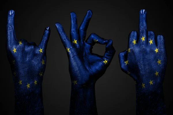 Un conjunto de tres manos con una bandera pintada de la UE muestran el dedo medio, cabra y bien, un signo de agresión, protesta y aprobación sobre un fondo oscuro . — Foto de Stock