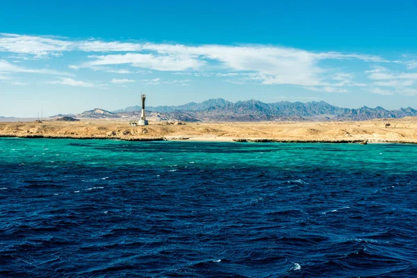 Морской пейзаж, высокий маяк на фоне синего моря с высокими лысыми горами на заднем плане — стоковое фото