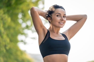 Bir spor üniforması koyu saçlı ince güzel kız bir gülümseme ile doğada gerilir ve çerçeve, sağlıklı bir yaşam tarzı ve atletik vücut sembolü içine bakmak.