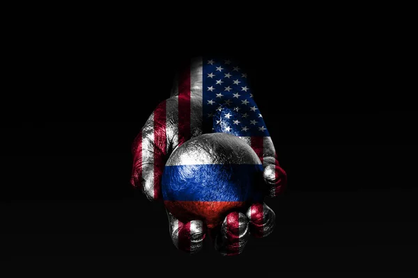 Uma mão com uma bandeira hasteada dos EUA segura uma bola com uma bandeira hasteada da Rússia, um sinal de influência, pressão ou conservação e proteção . — Fotografia de Stock