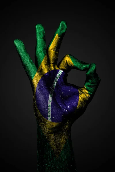 ブラジルの塗装された旗を持つ手は、暗い背景にOK記号を示しています. — ストック写真