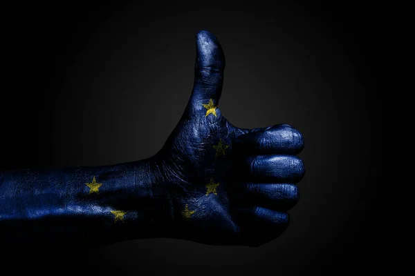 A hand with a drawn EU flag shows a finger up sign, a symbol of success, readiness, a task done on a dark background.