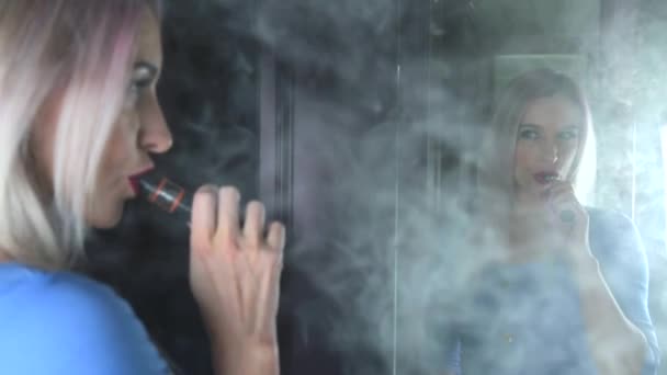 美丽的女人与粉红色的头发吸烟在镜子前的电子香烟 瓦平大量的烟雾 — 图库视频影像