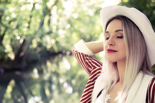 Una hermosa mujer rubia en un elegante traje con un sombrero blanco está de pie en un hermoso lugar natural . —  Fotos de Stock