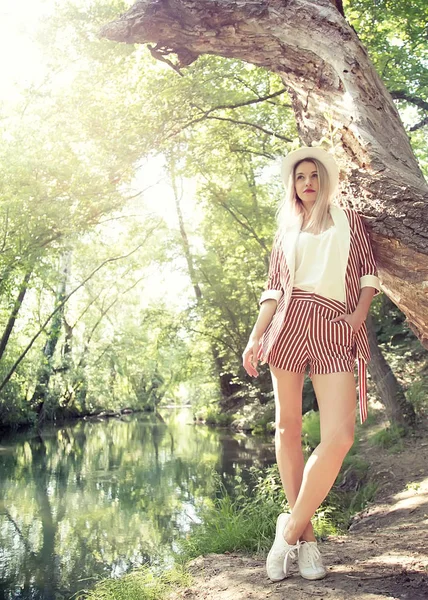 Una hermosa mujer rubia en un elegante traje con un sombrero blanco está de pie en un hermoso lugar natural . — Foto de Stock