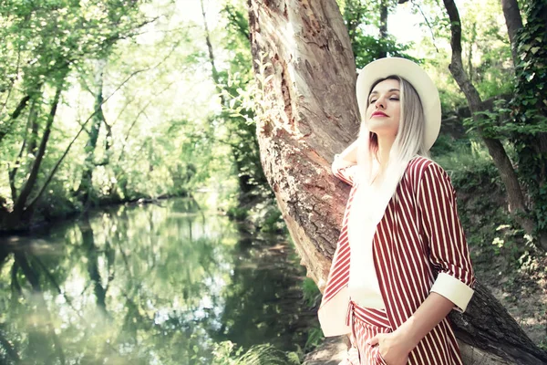 Una hermosa mujer rubia en un elegante traje con un sombrero blanco está de pie en un hermoso lugar natural . —  Fotos de Stock