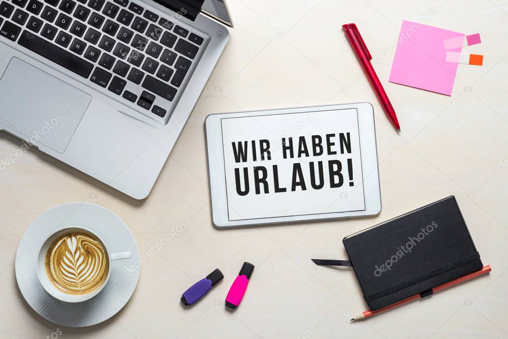 Wir haben Urlaub written in german on tablet meaning Closed for holidays as flatlay from above of an office desk background with notebook