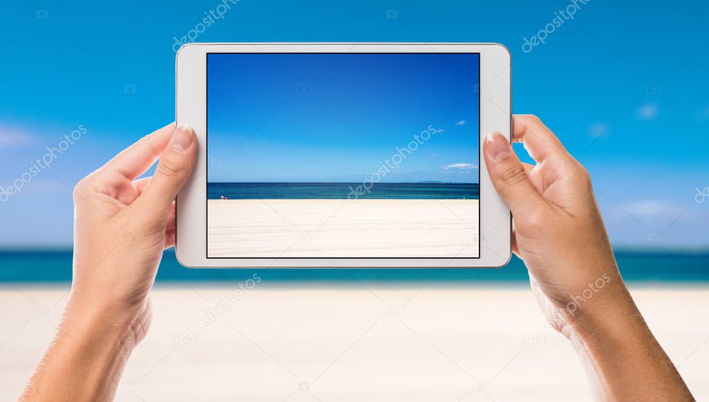 Holiday scene with beach and sea on white tablet screen with hands holding the device up against ocean and beach in the background 